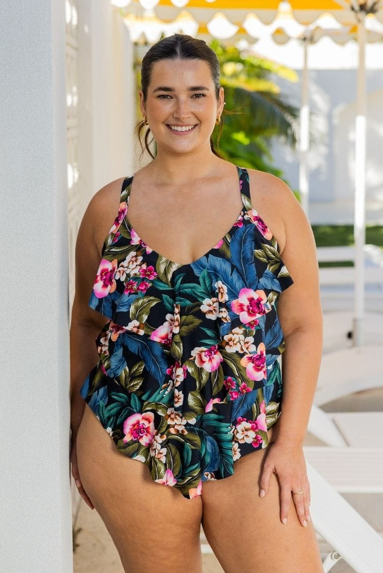A woman modeling a one-piece tropical floral swimsuit with a ruffled hem, standing in a bright, sunny outdoor setting with white lounge chairs and a yellow umbrella in the background.