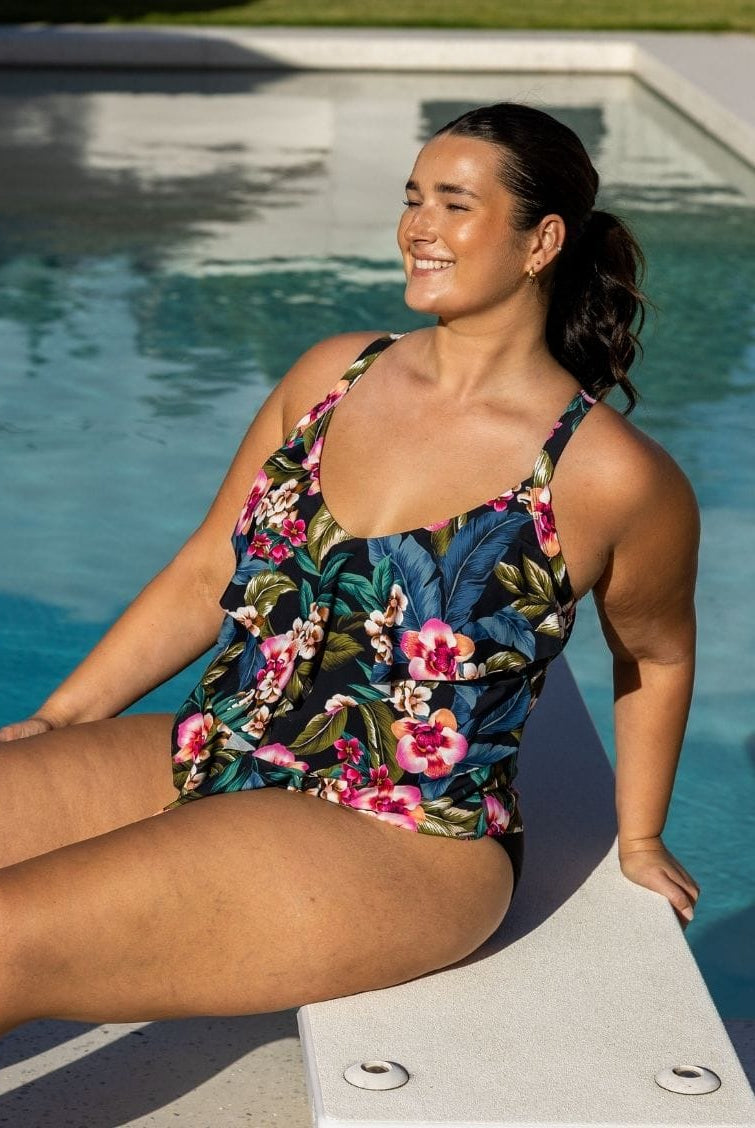 A woman wearing a floral one-piece swimsuit sits on the edge of a swimming pool outdoors.
