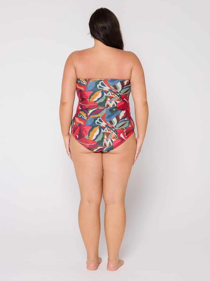 A woman viewed from behind wearing a strapless one-piece swimsuit with a colorful abstract print on a light gray studio background.