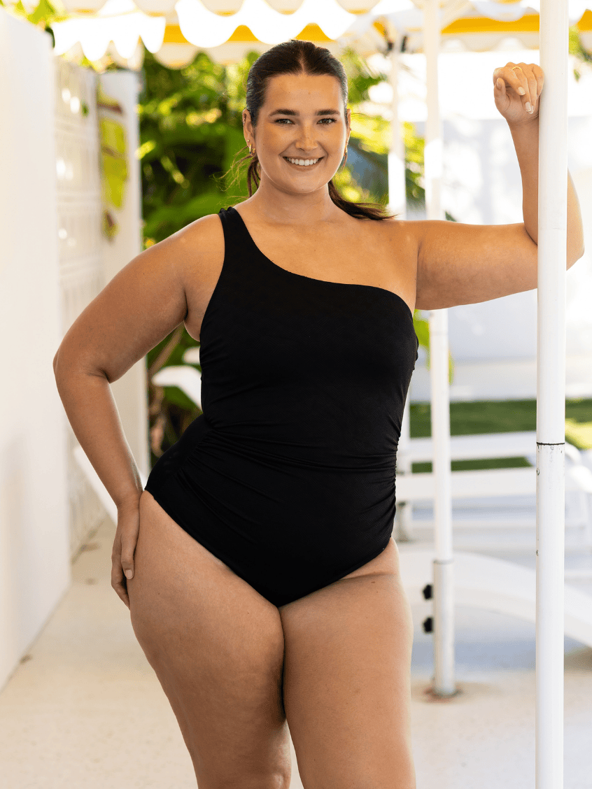 Woman wearing a black one-shoulder swimsuit standing outdoors.
