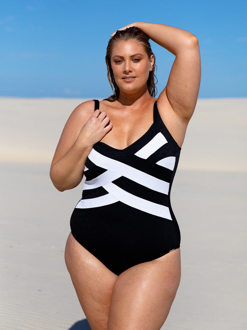 Woman wearing a black and white striped swimsuit against a blue sky and sand dunes background.