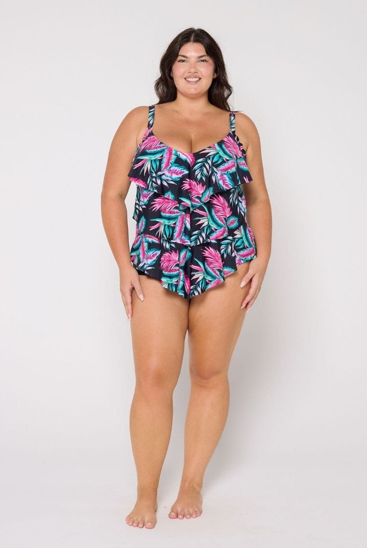 A woman modeling a 3 tier tankini ruffled swimsuit with a tropical palm leaf pattern in pink, teal, and black, standing against a plain white studio background.