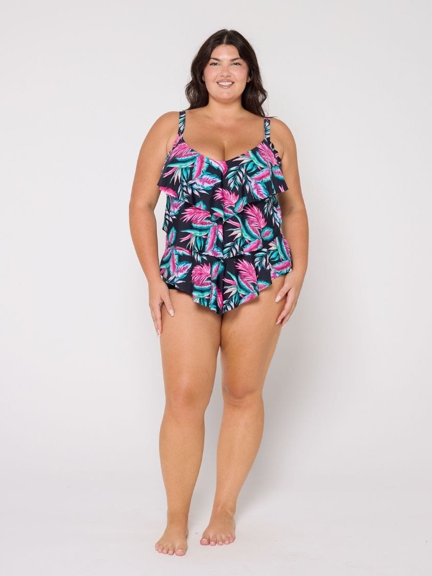 A woman modeling a 3 tier tankini ruffled swimsuit with a tropical palm leaf pattern in pink, teal, and black, standing against a plain white studio background.