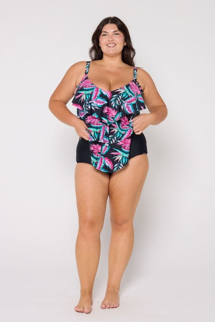 A woman modeling a one-piece swimsuit with a tropical leaf print on a black base, paired with solid black bottoms, against a plain light gray studio background.
