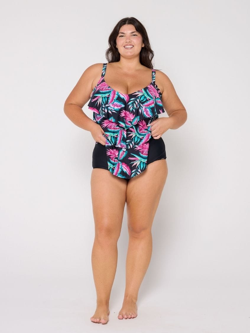 A woman modeling a one-piece swimsuit with a tropical leaf print on a black base, paired with solid black bottoms, against a plain light gray studio background.