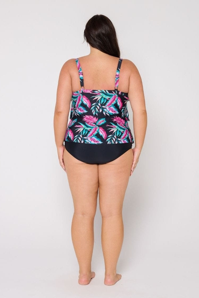 A woman viewed from the back wearing a 3 tiere tankini with a tropical leaf print on the top and solid black bottoms, standing against a light gray studio background.