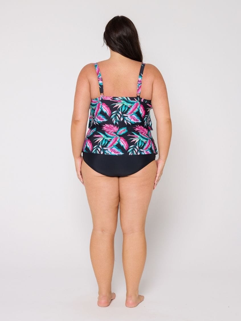 A woman viewed from the back wearing a 3 tiere tankini with a tropical leaf print on the top and solid black bottoms, standing against a light gray studio background.