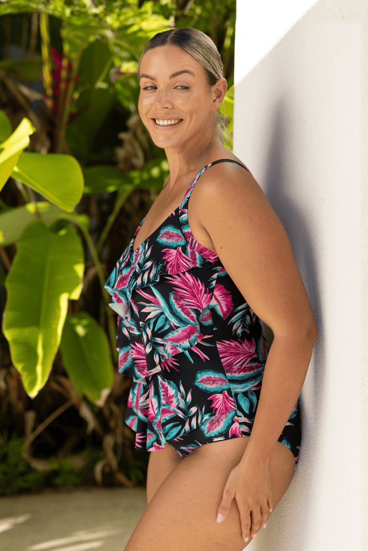 Woman wearing a tropical print swimsuit standing against a white wall with greenery in the background