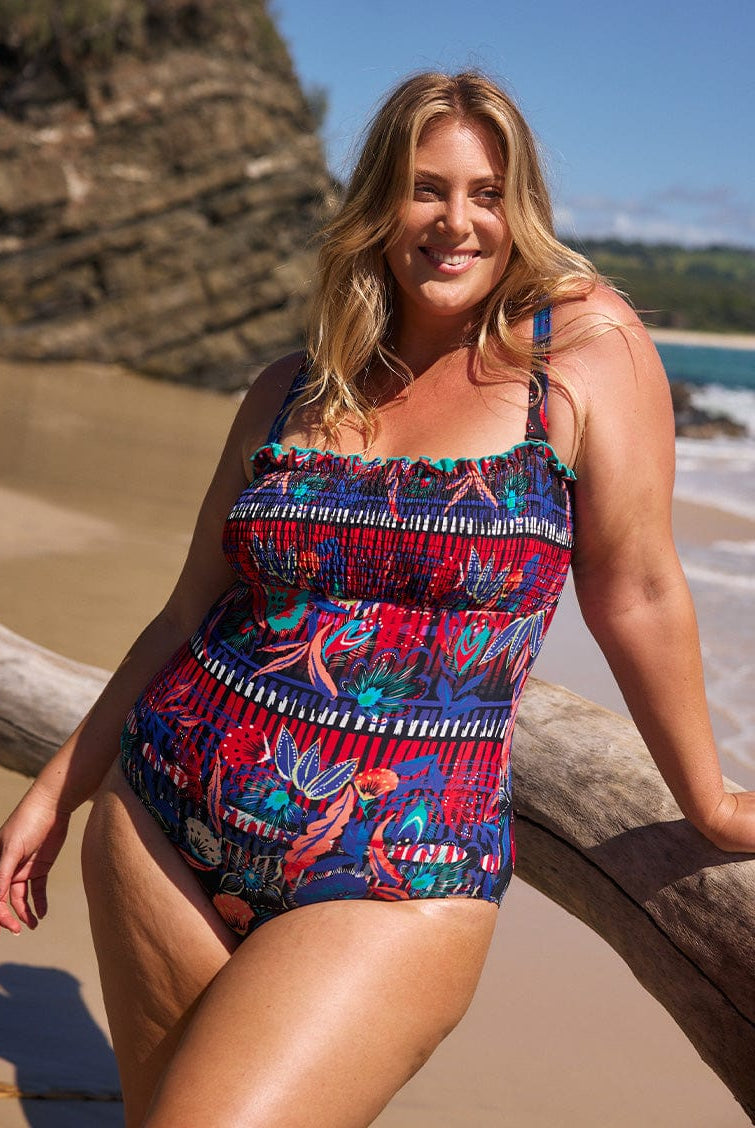 Blonde model on beach wearing a multi coloured pattern shirred one piece bandeau with straps for curve women