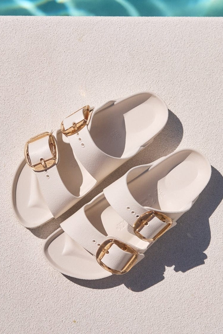 Pair of off white sandals with gold buckles on a diving board.