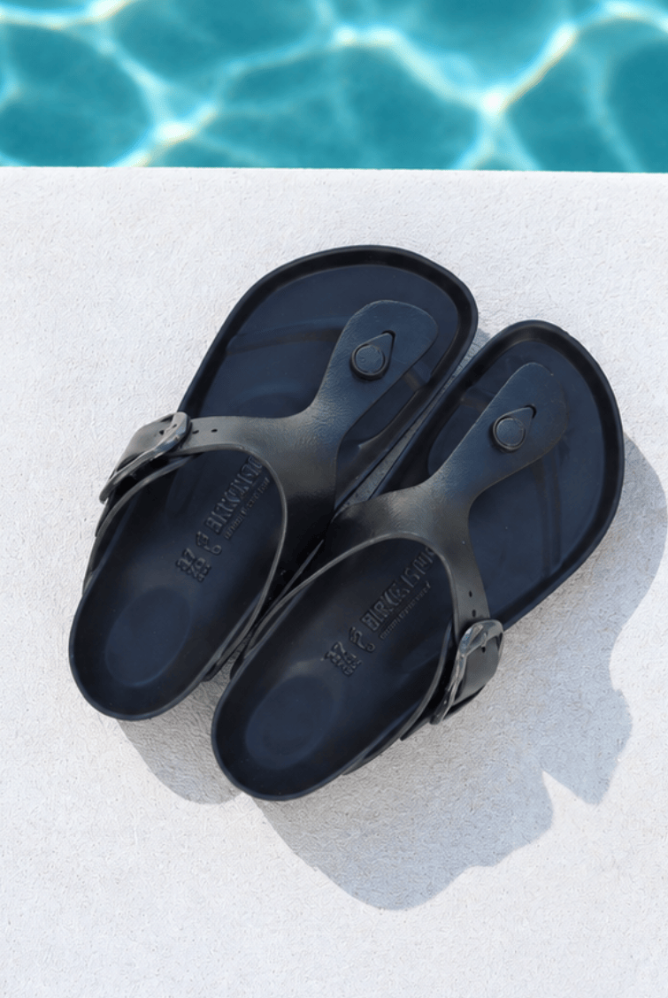 A pair of black rubber thong sandals with adjustable buckle straps, displayed on a light concrete surface with a swimming pool background.