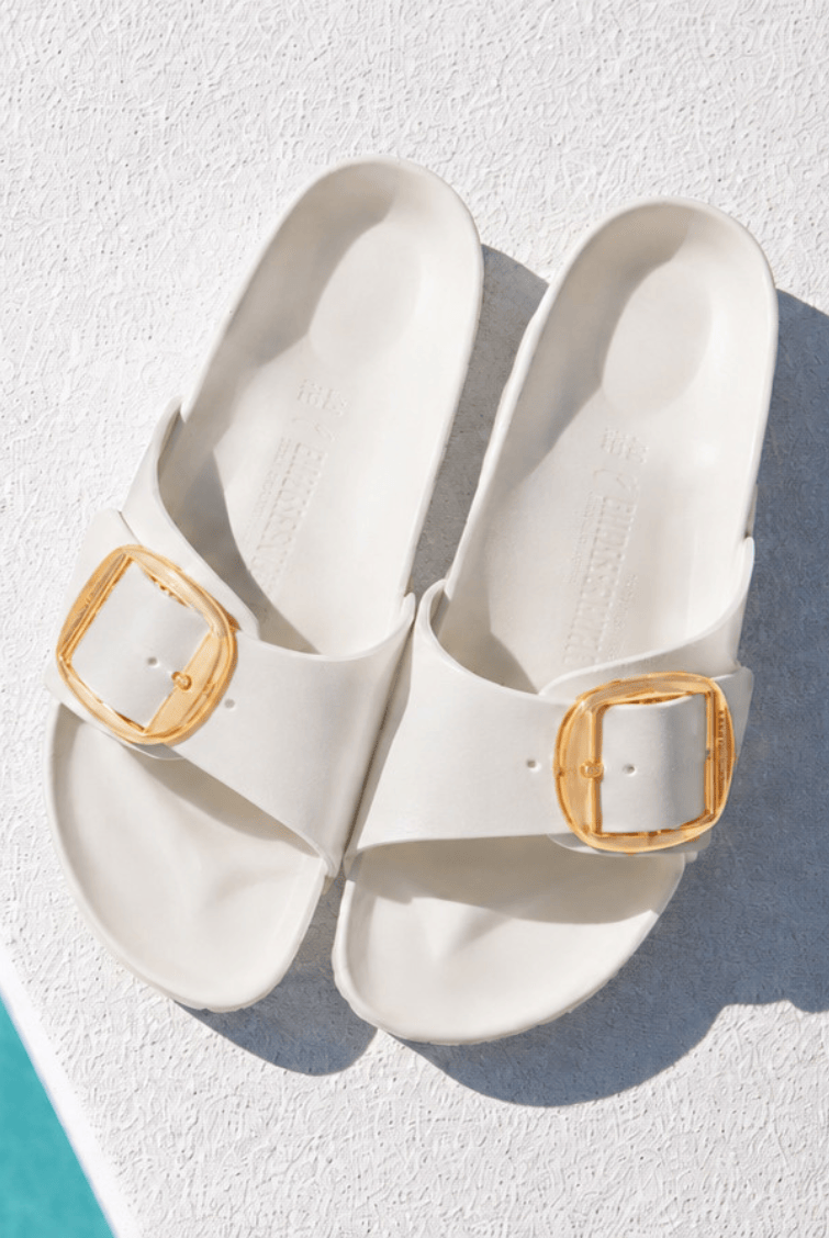A pair of off-white slide sandals with gold square buckle details, photographed from a top-down angle on a textured white surface with a turquoise edge visible in the bottom-left corner.