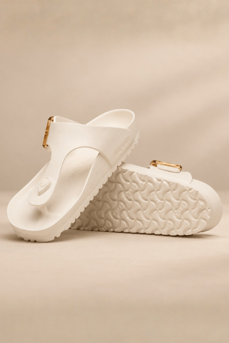A pair of white thong sandals with textured soles and small gold-tone buckle details, displayed on a warm beige background.