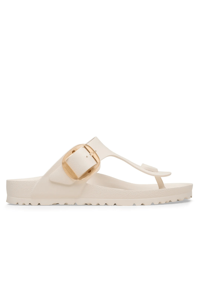 A single cream-colored thong sandal with a wide strap and a prominent gold buckle, shown in profile against a solid white background.