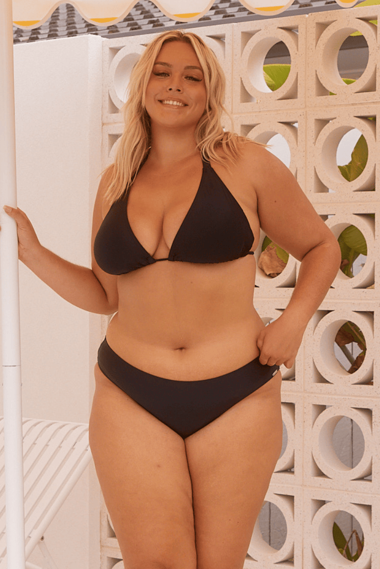 Woman wearing a black bikini with hipster pant standing against a decorative wall.