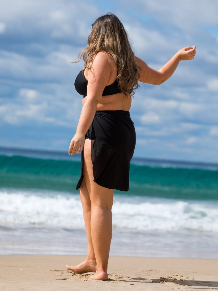 Woman in black swimsuit and sarong standing on a beach with ocean waves in the background