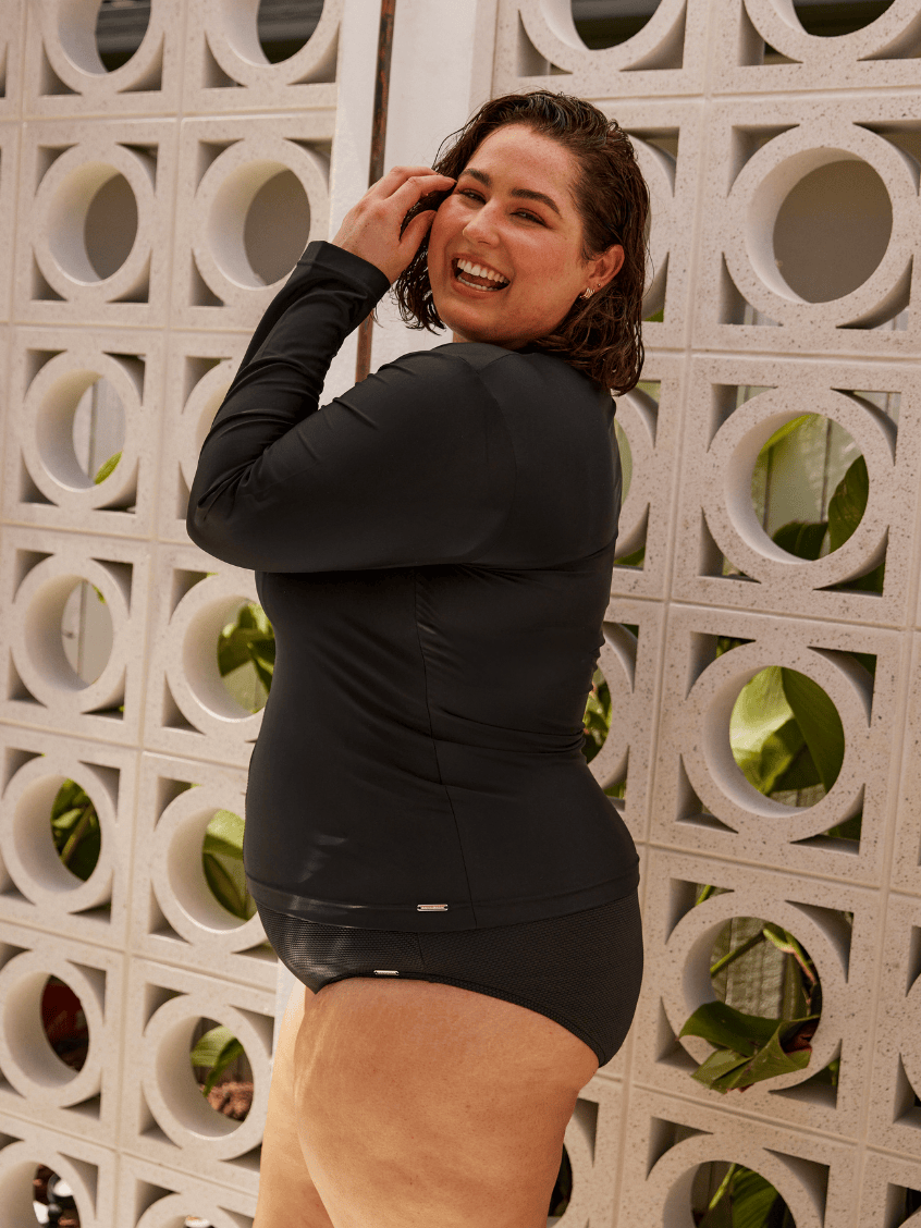 A woman wearing a black long-sleeve swim dress stands in front of a white concrete block wall with circular cutouts.