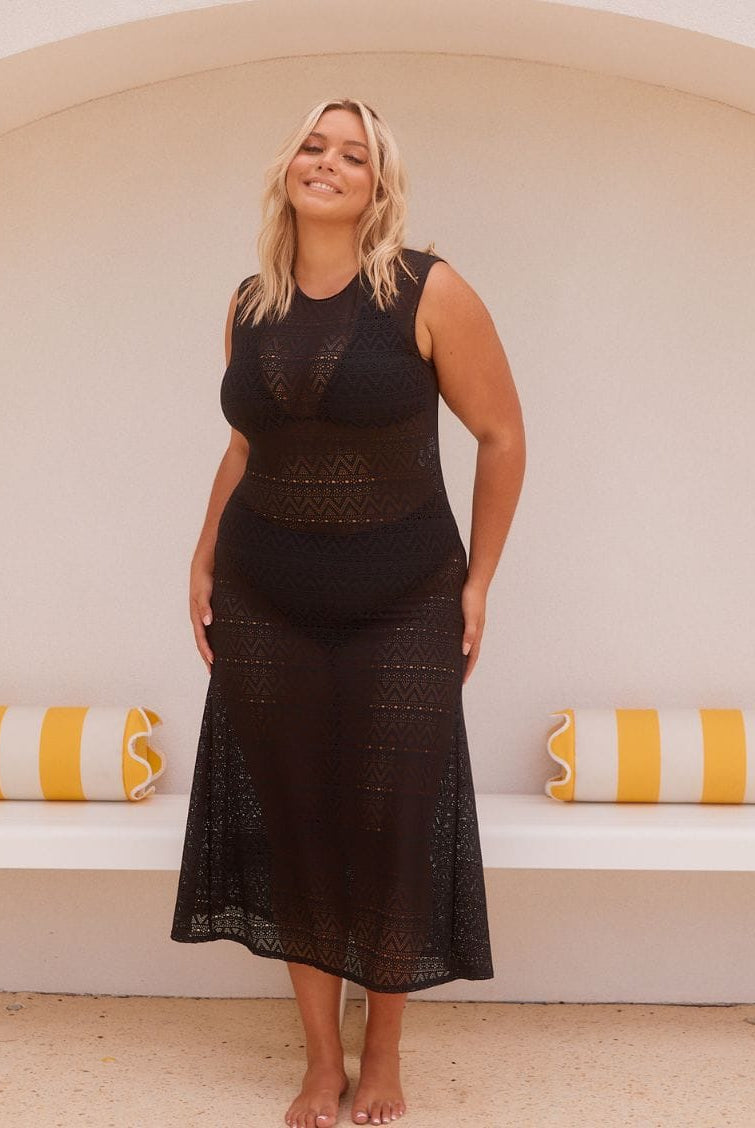 A woman modeling a black sleeveless crochet midi dress in front of a white arched wall with yellow and white striped pillows.
