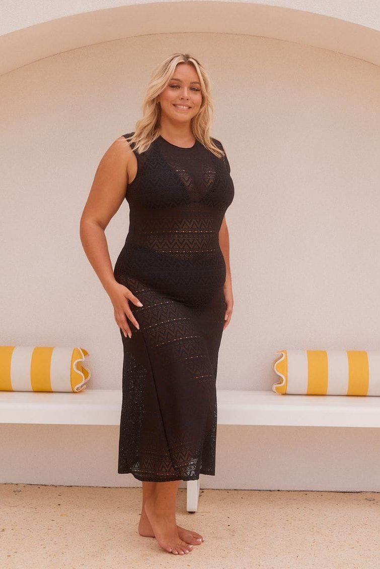 A woman stands barefoot in front of a white arched wall with yellow-and-white striped pillows, wearing a sleeveless black lace maxi dress.
