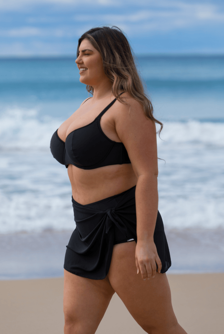 A woman walking on a sandy beach wearing a black two-piece swimwear set consisting of a bandeau-style bikini top and a matching wrap-style bikini skirt.