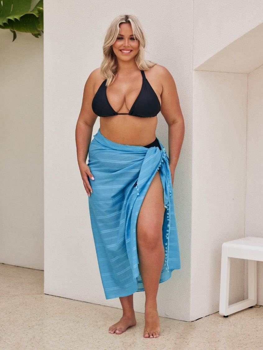 Woman wearing a black bikini top and blue sarong standing indoors.