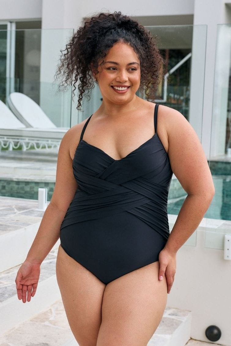 Brunette model wearing a black classic one piece with criss cross detail in metallic fabric