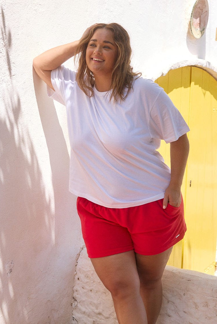 Curve woman wearing a white t-shirt and red shorts standing against a white wall with a yellow archway.