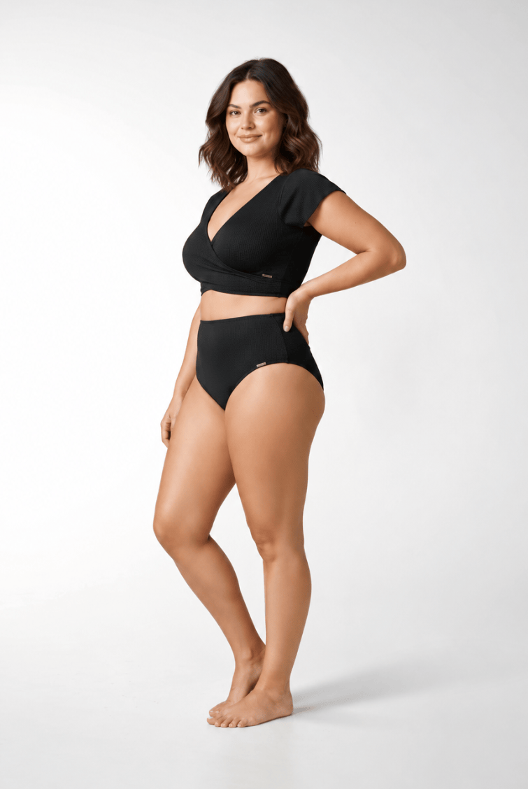 A woman modeling a black two-piece swimsuit consisting of a short-sleeve wrap-style bikini top and matching high-waisted bikini bottoms, photographed against a clean light gray studio background.