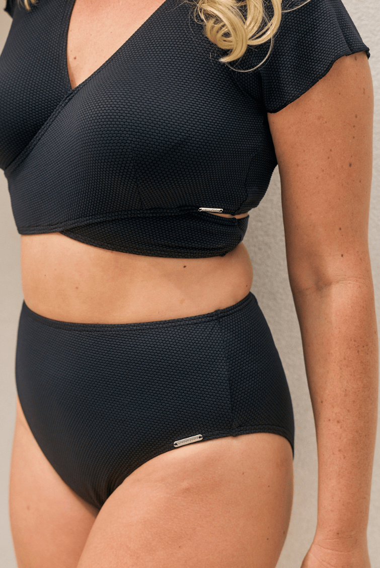 A woman modeling a black two-piece swimsuit consisting of a short-sleeve wrap-front crop top and matching high-waisted briefs, photographed against a seamless light gray studio background.