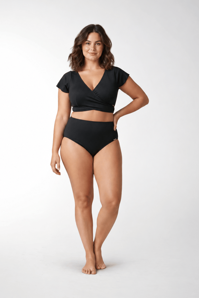 A woman modeling a black two-piece swimsuit consisting of a short-sleeve wrap-front crop top and matching high-waisted briefs, photographed against a seamless light gray studio background.