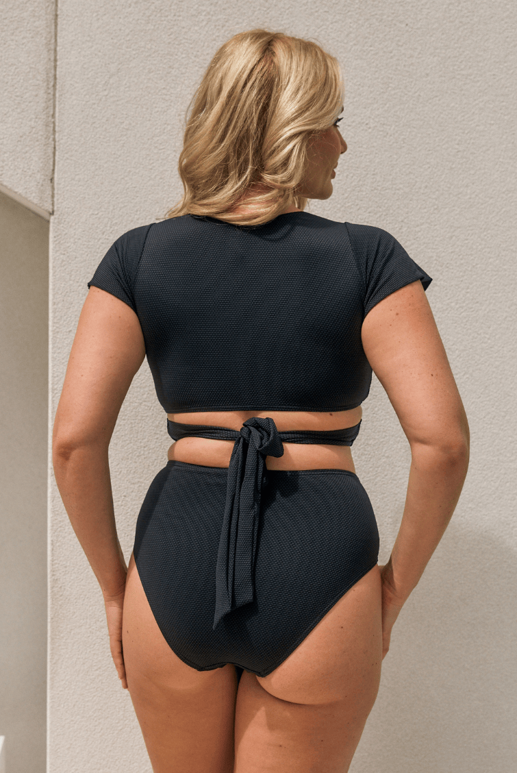 A woman shown from the back wearing a black two-piece swimsuit with a short-sleeve crop top and high-waisted ribbed bottoms tied at the waist, standing against a light beige textured wall.