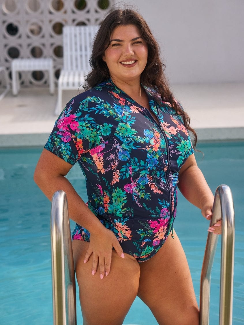 Woman in a colorful floral rash vest swimsuit standing by a pool.