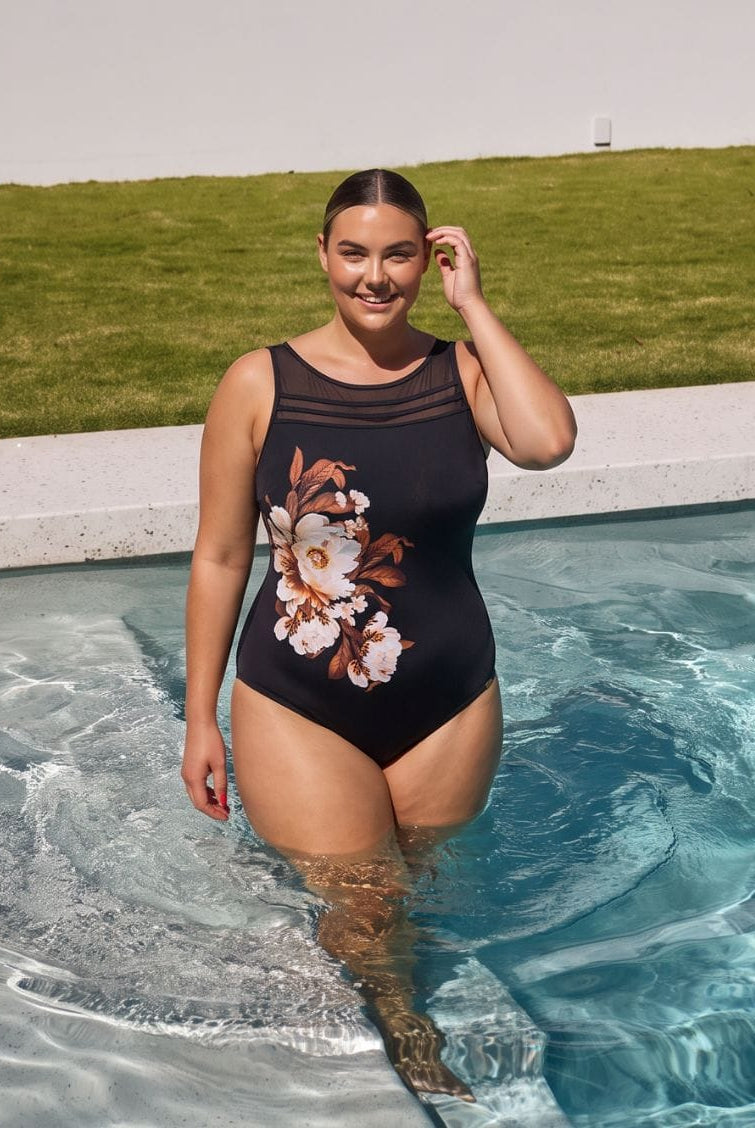 A woman stands in a swimming pool wearing a black one-piece swimsuit with a sheer mesh neckline and a large white and brown floral print on the front. A grassy lawn is visible in the background under a bright sky.