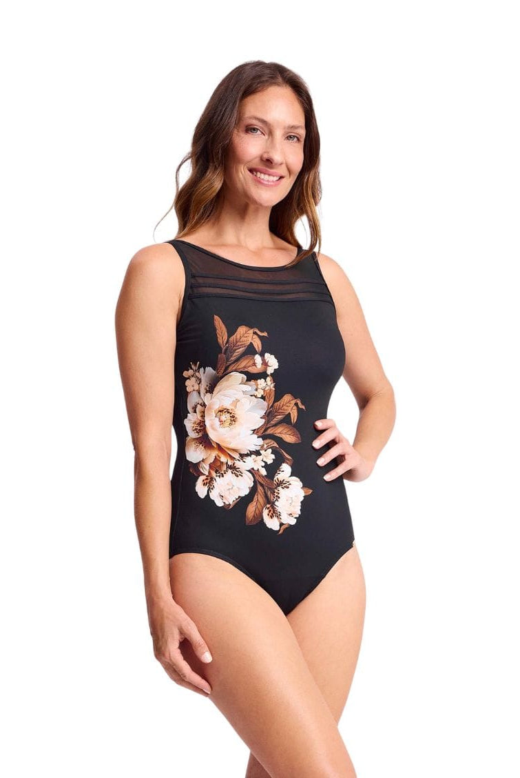 A woman modeling a black sleeveless one-piece swimsuit with a sheer mesh chest panel and a large floral print in cream, white, and brown on the front, set against a plain white background.