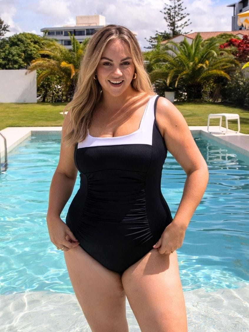 Woman in a black and white square neck swimsuit standing by a pool with greenery in the background