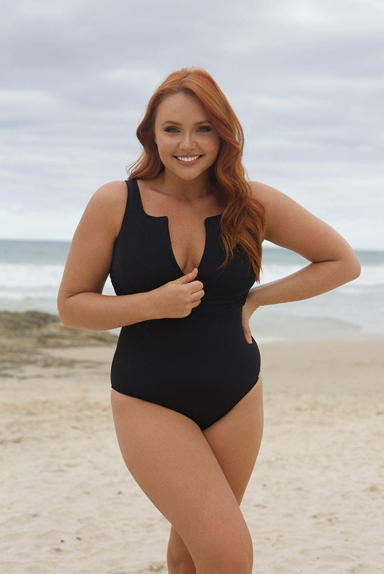 Woman in a black swimsuit standing on a beach with ocean and sky in the background