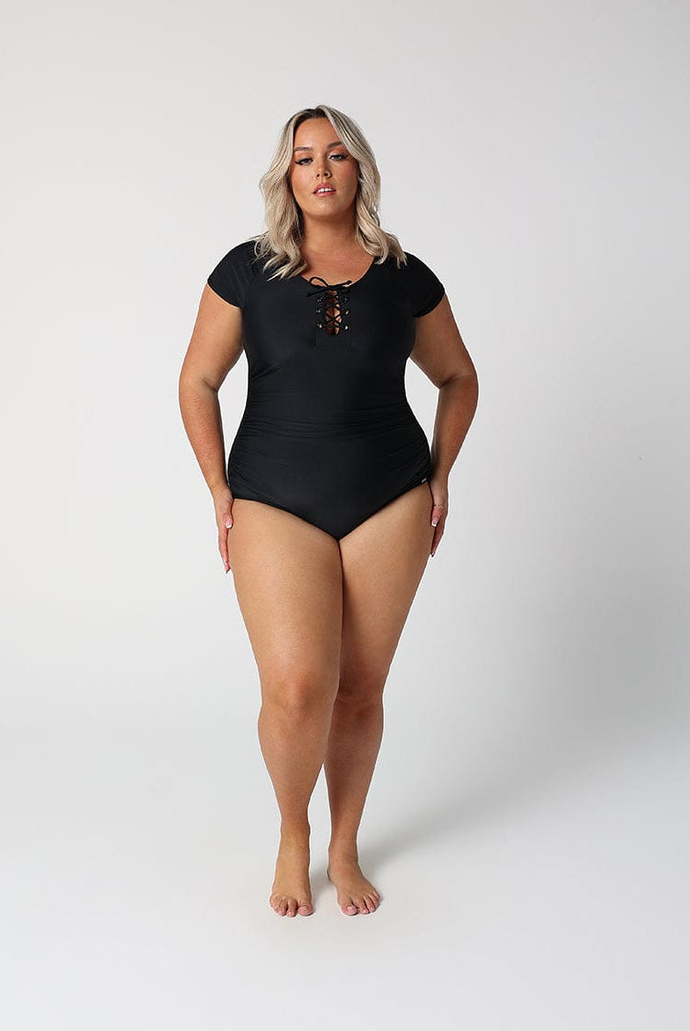 Woman wearing a black swimsuit with a lace-up front, standing in front of a white wall