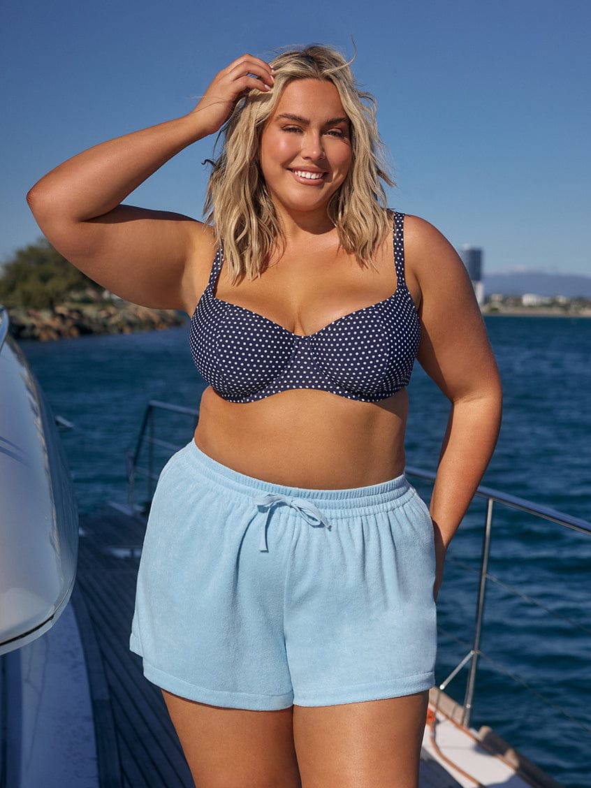 Woman in a navy bikini top and light blue shorts standing on a boat with water and sky in the background.