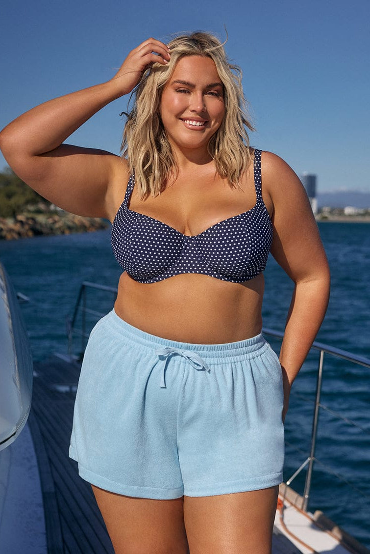 Woman in a navy bikini top and light blue shorts standing on a boat with water and sky in the background.