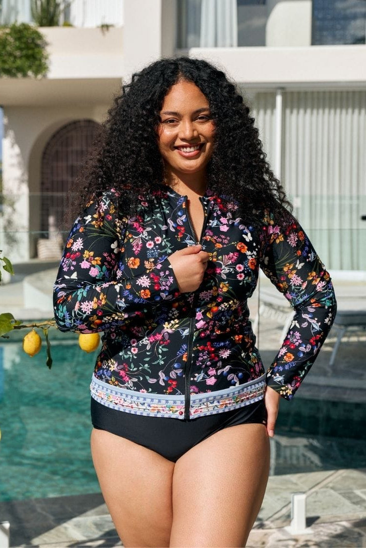 A woman wearing a floral long-sleeve swim jacket over a black bikini stands by a swimming pool in front of a modern white building.