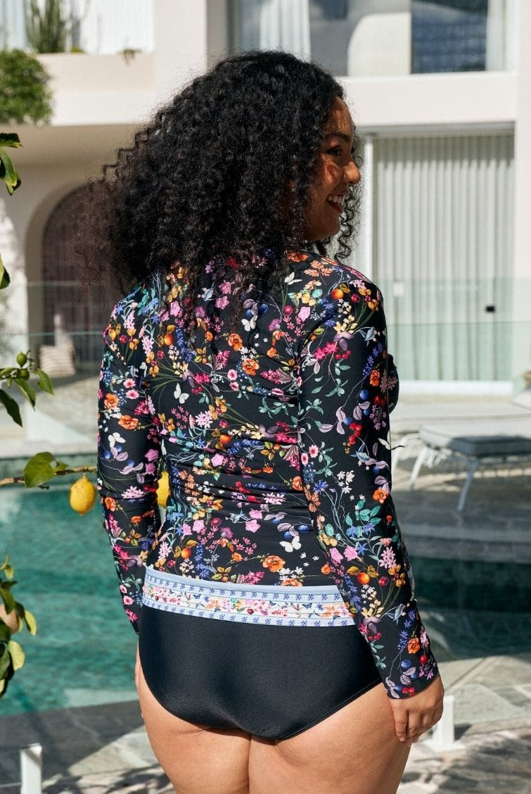 A woman standing by a swimming pool wearing a long-sleeve floral swim cover-up with black high-waisted bottoms. The cover-up features a multicolor floral pattern on a black background with a decorative multicolor border at the waist.