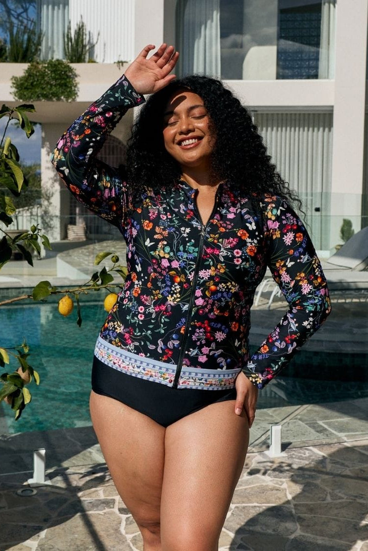 A woman posing by a swimming pool wearing a long-sleeve floral swim cover-up top with black swim briefs.