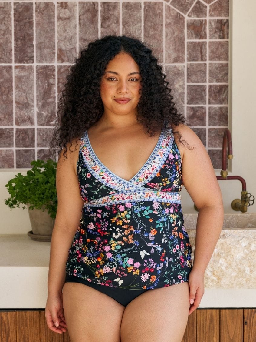 A woman modeling a floral print one-piece swimsuit with a blue and white patterned trim, standing in front of a tiled wall and sink.