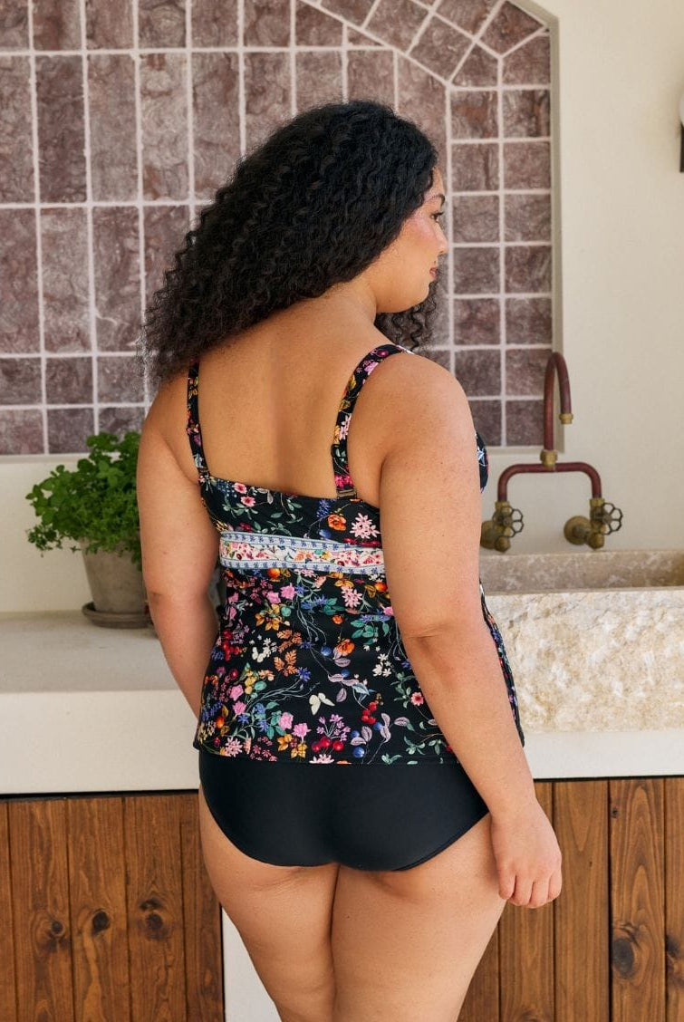 A woman shown from the back wearing a floral print tankini swimsuit top and solid black bottoms, standing in front of a kitchen sink with tiled backsplash.