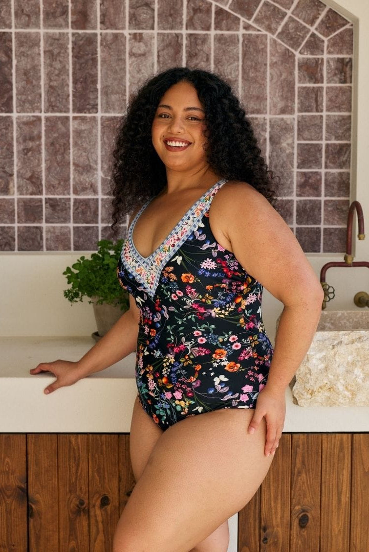 A woman modeling a floral one-piece swimsuit in a kitchen setting with a tiled backsplash and wooden cabinets.