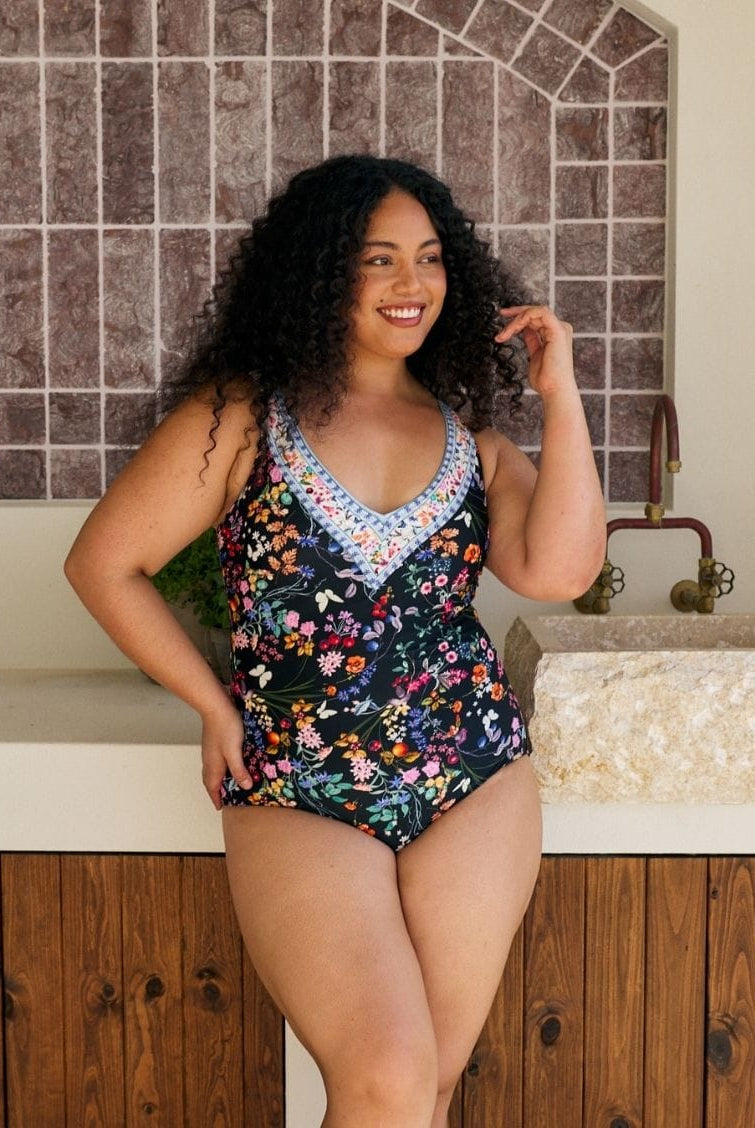 A woman modeling a one-piece swimsuit with a dark navy floral print and a light blue crochet V-neck trim, standing in front of a tiled wall and stone sink.