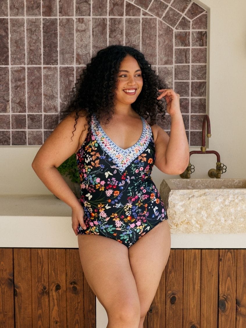 A woman modeling a one-piece swimsuit with a dark navy floral print and a light blue crochet V-neck trim, standing in front of a tiled wall and stone sink.