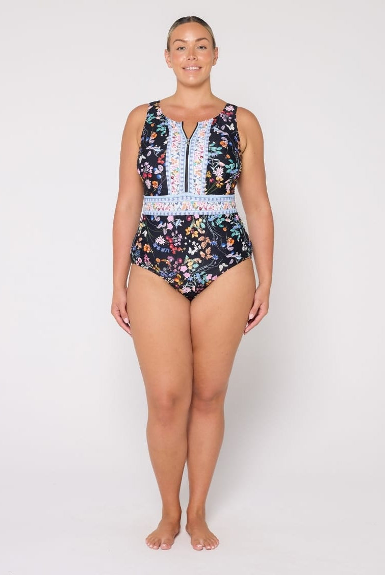 A woman modeling a sleeveless one-piece swimsuit with a dark navy floral print, a white patterned waistband, and a central zipper, photographed against a plain light gray studio background.