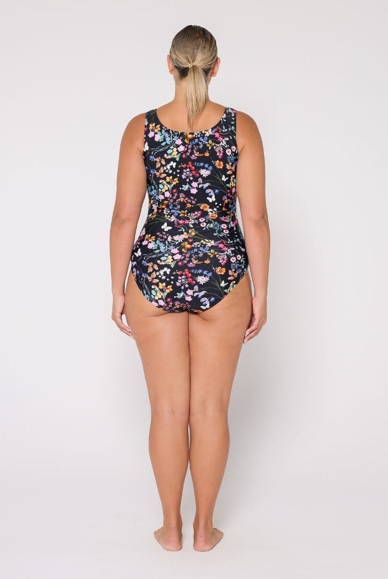 A woman viewed from the back wearing a sleeveless one-piece swimsuit with a dense multicolor floral print on a black background, standing against a light gray studio backdrop.