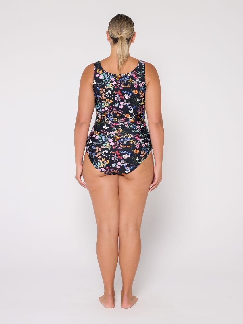 A woman viewed from the back wearing a sleeveless one-piece swimsuit with a dense multicolor floral print on a black background, standing against a light gray studio backdrop.